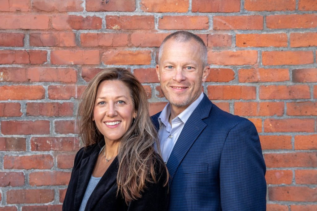 professional-man-and-woman-14634936 A cheerful middle-aged man and woman in business attire posing against a brick wall, depicting professionalism and partnership.